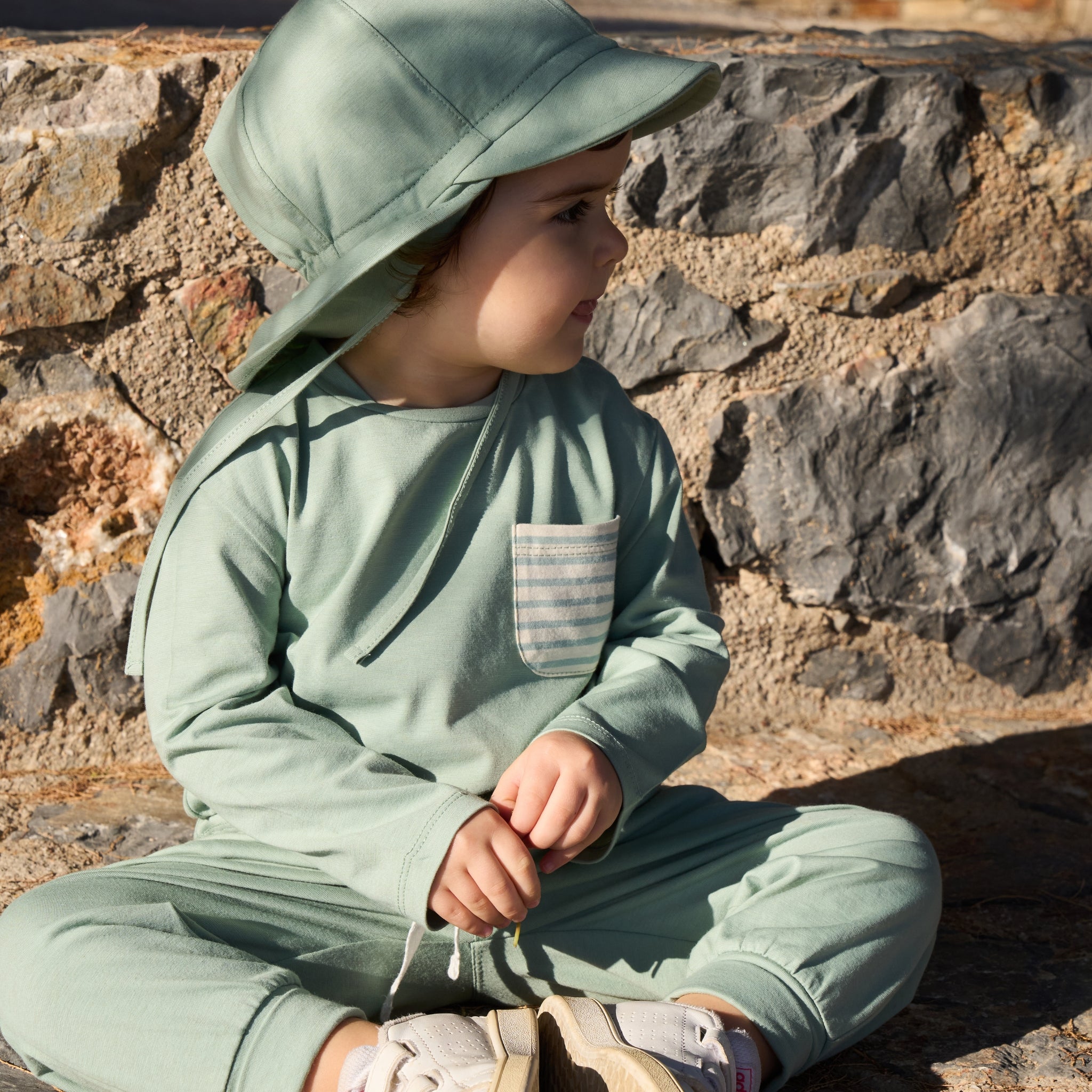 Baby sitting on a stone wall wearing a UPF50+ Pants, UPF+ Long sleeve and UPF50+ Sunhat.