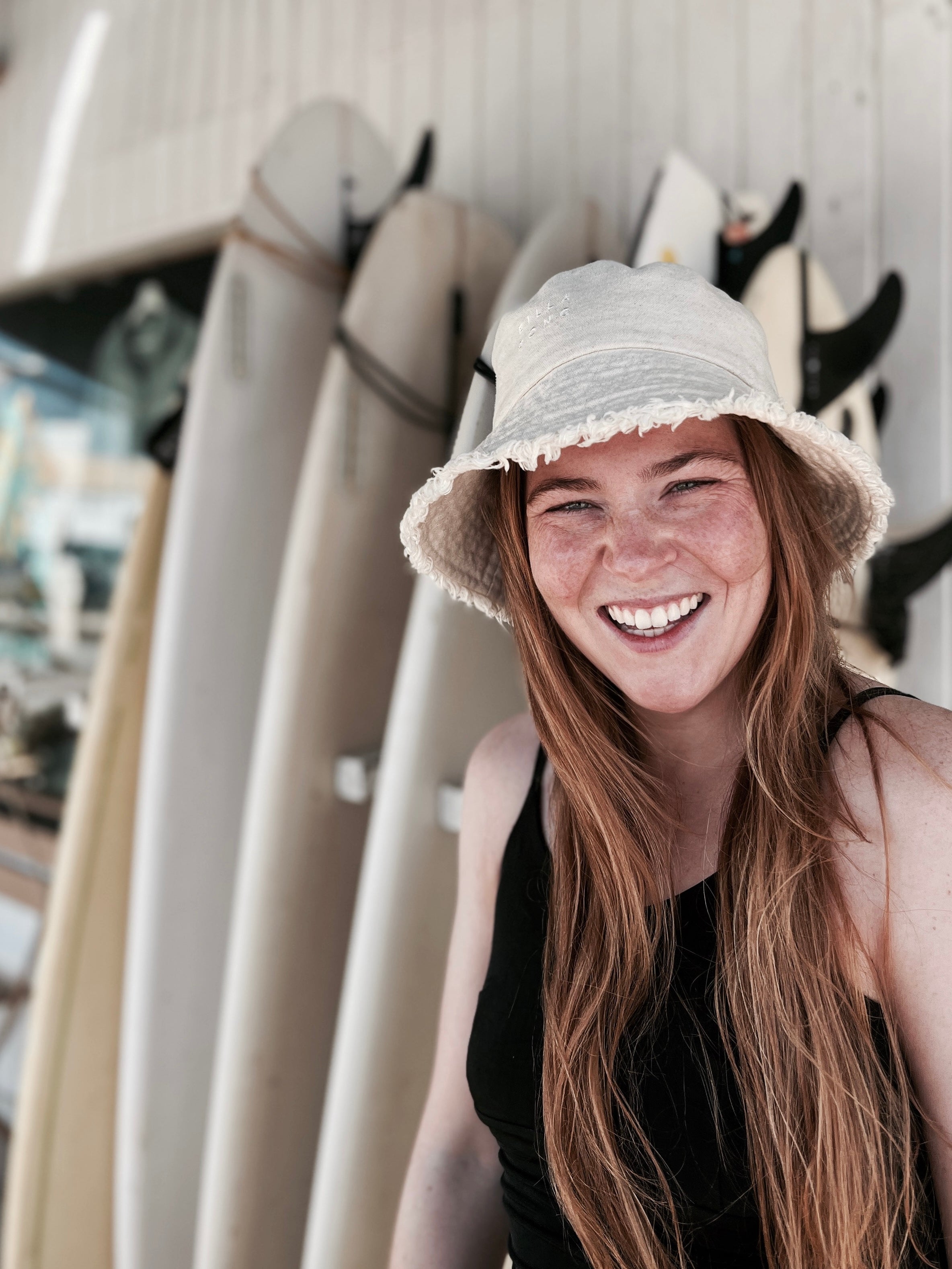 Bild der Gründerin mit einem Hut und Surfbrettern im Hintergrund.