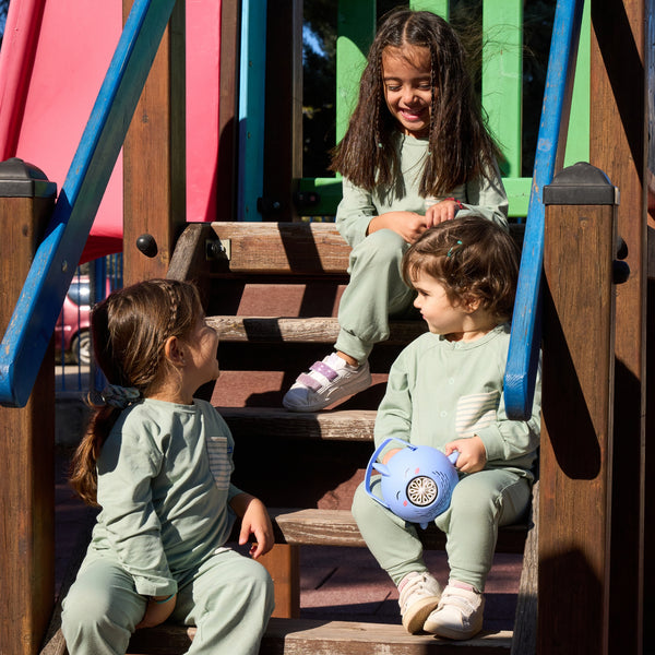 Drei Kinder sitzen auf einer Spielplatz-Treppe und lachen sich an. Sie tragen alle UV-Schutz Kleidung für Babys und Kinder: UV Schutz Hose, UV Schutz Shirt und ein UV Schutz Einteiler.