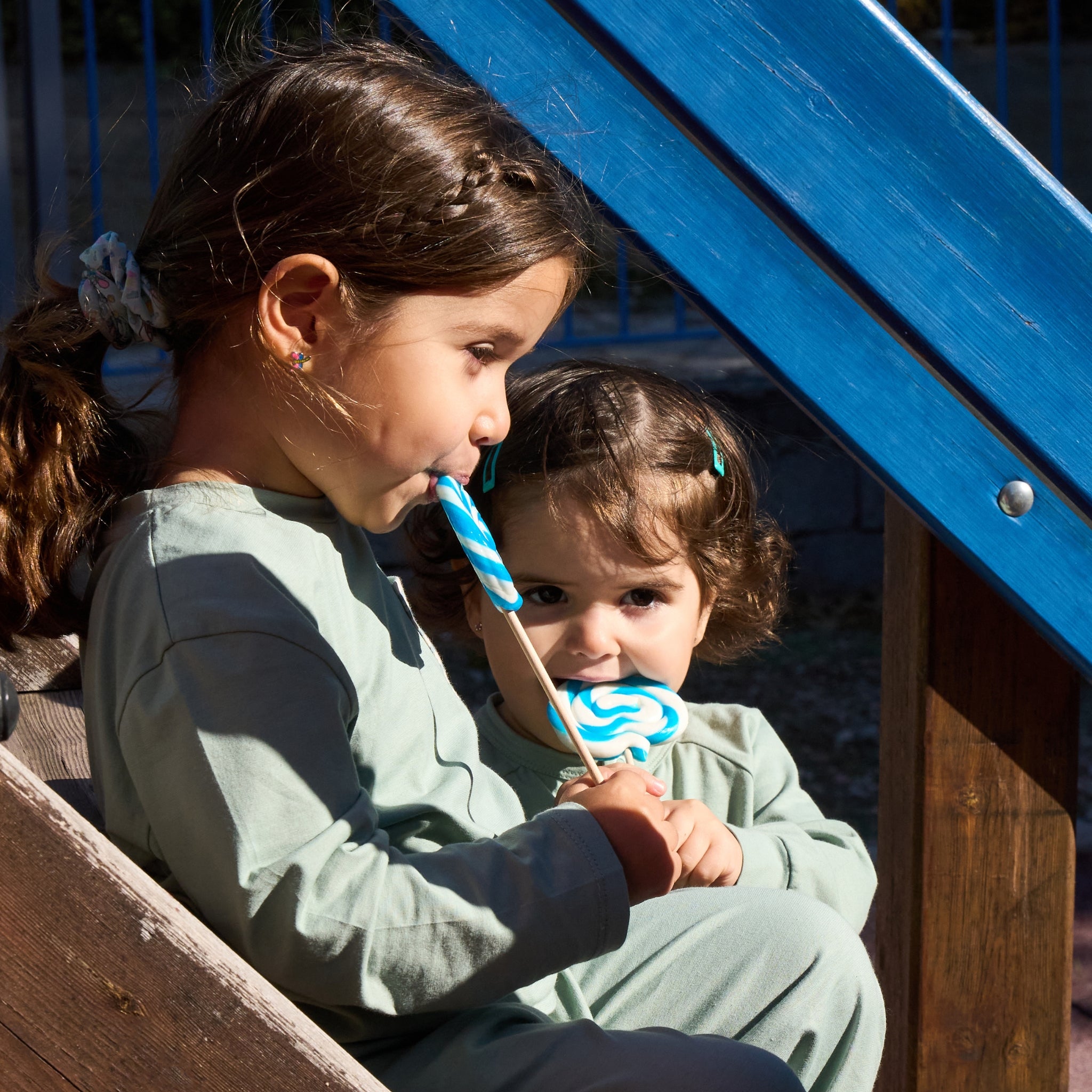 Zwei Kinder, die UV-Schutzkleidung tragen, auf einer Holztreppe sitzen und einen bunten Lolli essen.