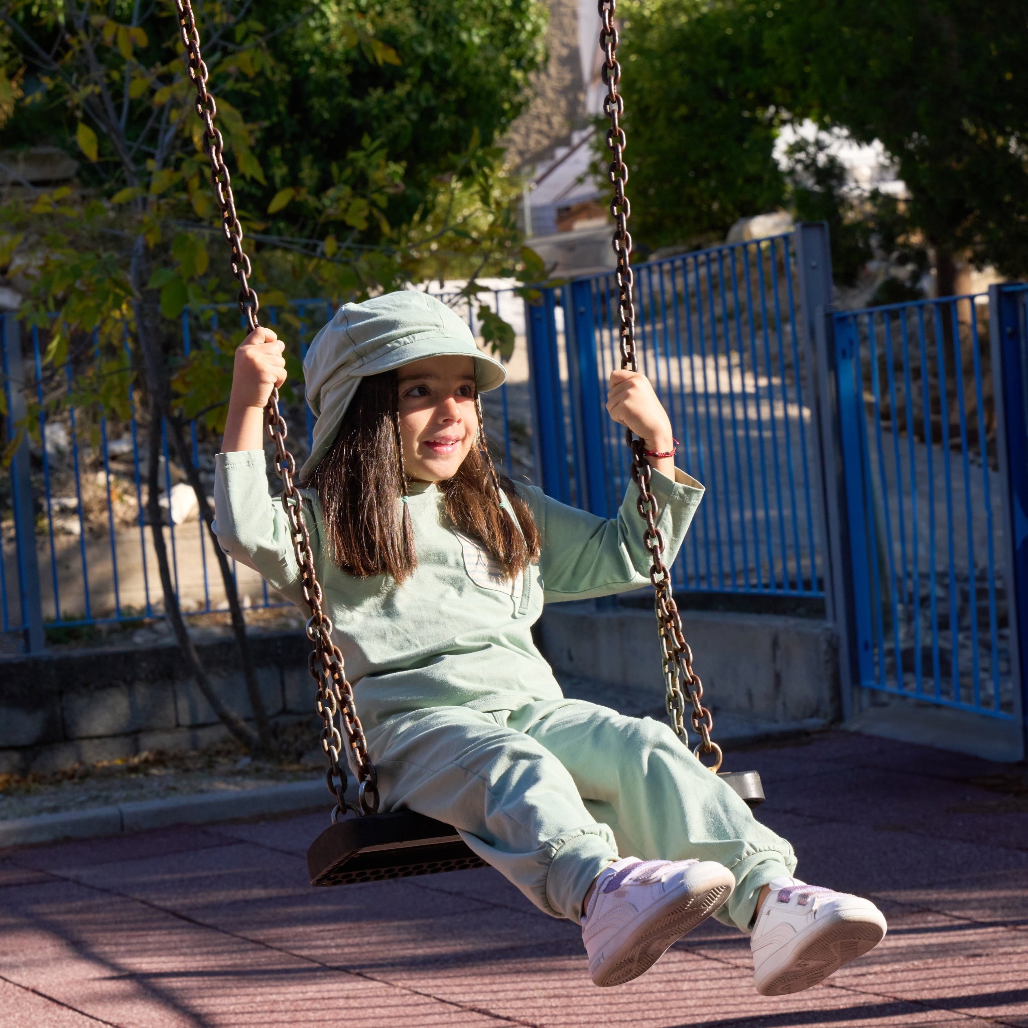 Ein Mädchen sitzt auf einer Schaukel und trägt eine UV-Schutz Hose, ein UV-Schutz Shirt und ein UV-Schutz Hut mit Kappe und Nackenschutz.