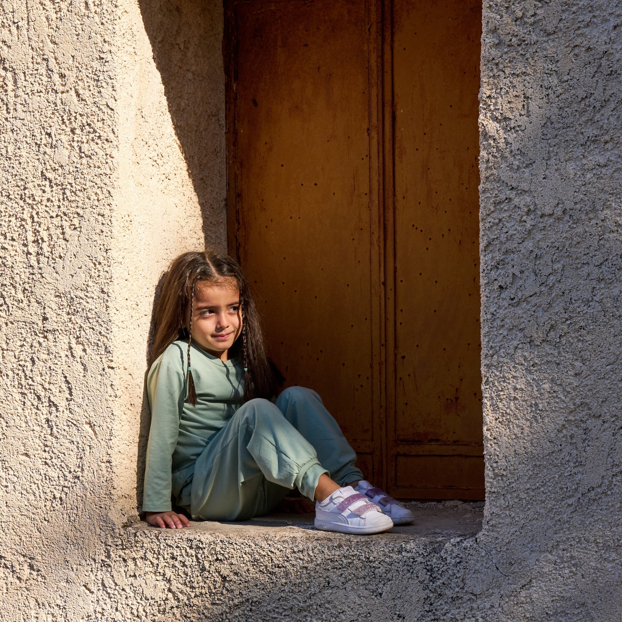 Ein Mädchen sitzt in einer Ecke eines Fensters in der Sonne und trägt Sonnenschutzkleidung für Kinder mit UPF50+.